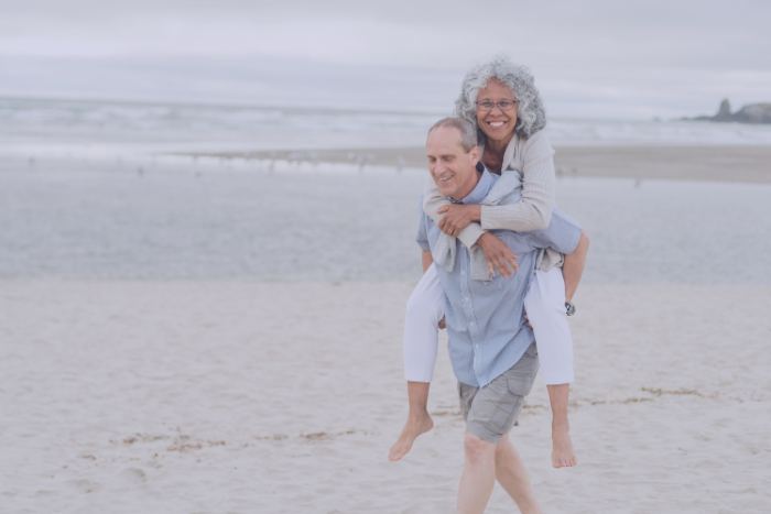 Joint replacement can be life-changing Older couple on beach with man giving woman a piggy back